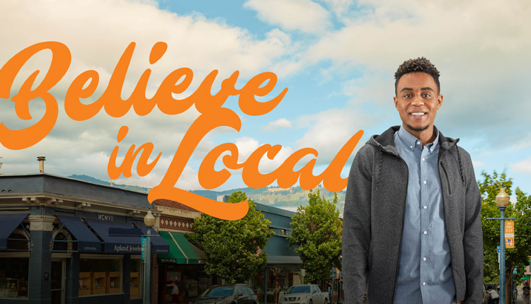 A young man smiles as he stands in front of a downtown scene, flanked by the Believe in Local logo.